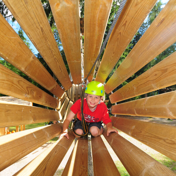 Ein kleines Kind, das einen Helm und einen Klettergurt trägt, krabbelt an einem sonnigen Tag durch eine hölzerne Tunnelstruktur in einem Outdoor-Abenteuerpark.