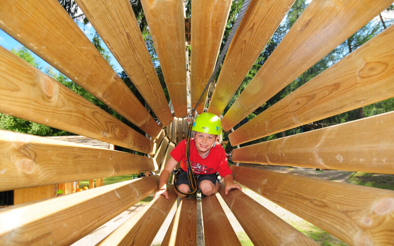 Ein kleines Kind, das einen Helm und einen Klettergurt trägt, krabbelt an einem sonnigen Tag durch eine hölzerne Tunnelstruktur in einem Outdoor-Abenteuerpark.