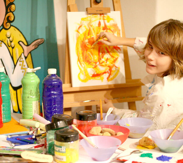 Ein Kind malt mit gelben und roten Farben auf einer Leinwand an einem Tisch voller Künstlerbedarf, darunter Farbflaschen, Pinsel und Schüsseln.