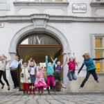 Eine Gruppe von Kindern springt in die Luft vor einem historischen Gebäude mit gewölbtem Eingang und dem Schild "Dom Gasse".