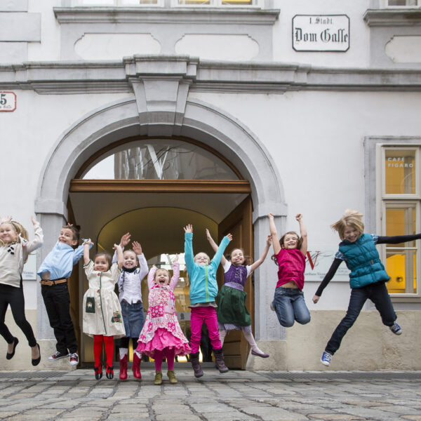 Eine Gruppe von Kindern springt in die Luft vor einem historischen Gebäude mit gewölbtem Eingang und dem Schild "Dom Gasse".