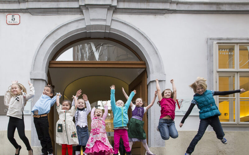 Eine Gruppe von Kindern springt in die Luft vor einem historischen Gebäude mit gewölbtem Eingang und dem Schild "Dom Gasse".