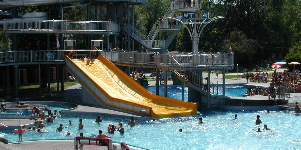 Menschen schwimmen in einem öffentlichen Schwimmbad und nutzen eine große gelbe Wasserrutsche, während andere von Bänken und schattigen Bereichen in der Nähe aus zuschauen.