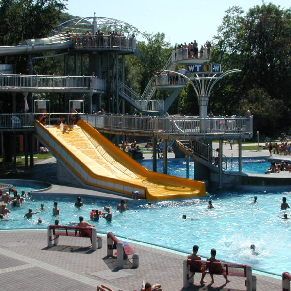 Menschen schwimmen in einem öffentlichen Schwimmbad und nutzen eine große gelbe Wasserrutsche, während andere von Bänken und schattigen Bereichen in der Nähe aus zuschauen.