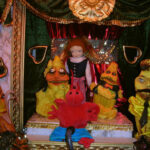 Auf einer Puppenbühne sind eine Puppe in karierten Hosen, eine rote Krabbenpuppe und vier gelbe Puppen mit unterschiedlichen Gesichtern zu sehen, alle vor einem kunstvollen Hintergrund mit grünen und roten Vorhängen.