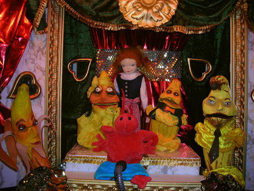 Auf einer Puppenbühne sind eine Puppe in karierten Hosen, eine rote Krabbenpuppe und vier gelbe Puppen mit unterschiedlichen Gesichtern zu sehen, alle vor einem kunstvollen Hintergrund mit grünen und roten Vorhängen.