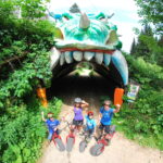 Vier Kinder mit Fahrrädern und Helmen posieren vor einem Tunneleingang, der wie ein Drachenmaul aussieht, umgeben von Grünpflanzen.