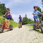 Drei Personen mit Helmen fahren an einem sonnigen Tag auf einem von Gras und Bäumen umgebenen Kiesweg mit einem Geländemotorroller.