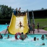 Kinder spielen in einem Freibad, einige schwimmen, während andere eine gelbe Wasserrutsche benutzen; die Szene ist lebhaft und spielt sich an einem sonnigen Tag ab.