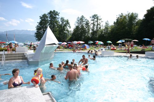 Menschen verschiedenen Alters schwimmen und entspannen sich in einem Außenpool mit Sonnenschirmen, Bäumen und Bergen im Hintergrund.
