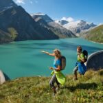 Zwei Wanderer mit Rucksäcken stehen auf einem grasbewachsenen Hang mit Blick auf einen türkisfarbenen See, der von Bergen und einem großen Staudamm begrenzt wird, unter einem klaren blauen Himmel.