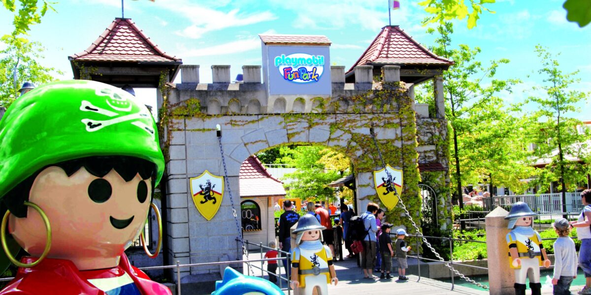 Eingang zum Playmobil FunPark mit großen Playmobil-Figurenstatuen, einem burgähnlichen Tor und Besuchern, die an einem sonnigen Tag eintreten.