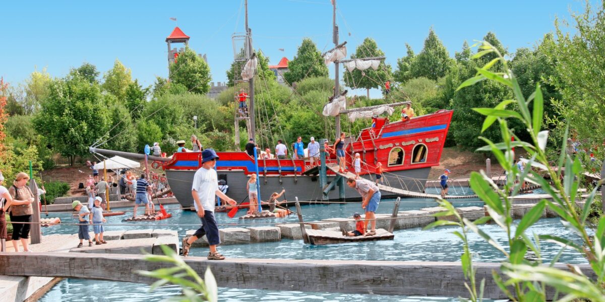 Menschen laufen auf Holzbrücken und Flößen über einen Teich in einem Park im Freien mit einem großen roten Piratenschiff und grünen Bäumen im Hintergrund.