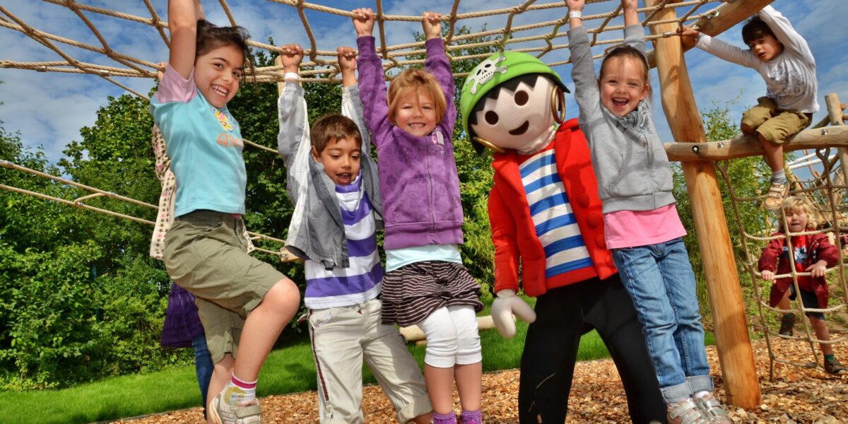 Fünf Kinder und eine Person im Kostüm einer Zeichentrickfigur hängen lächelnd an einem Seilklettergerüst auf einem Außenspielplatz mit Holzspänen und Grünpflanzen.