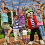 Fünf Kinder und eine Person im Kostüm einer Zeichentrickfigur hängen lächelnd an einem Seilklettergerüst auf einem Außenspielplatz mit Holzspänen und Grünpflanzen.