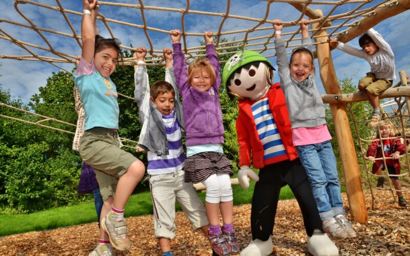 Fünf Kinder und eine Person im Kostüm einer Zeichentrickfigur hängen lächelnd an einem Seilklettergerüst auf einem Außenspielplatz mit Holzspänen und Grünpflanzen.