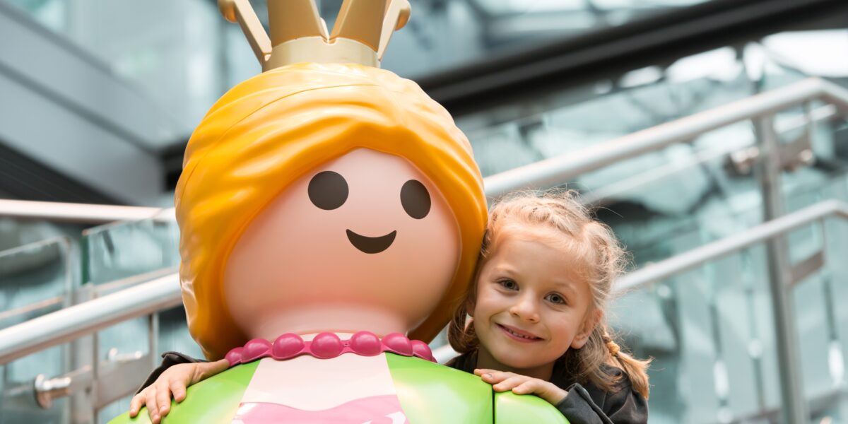 Ein junges Mädchen lächelt und umarmt eine große Playmobil-Prinzessinnenfigur mit einer Krone, drinnen in der Nähe von Glasfenstern und Metallgeländern.