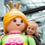 Ein junges Mädchen lächelt und umarmt eine große Playmobil-Prinzessinnenfigur mit einer Krone, drinnen in der Nähe von Glasfenstern und Metallgeländern.