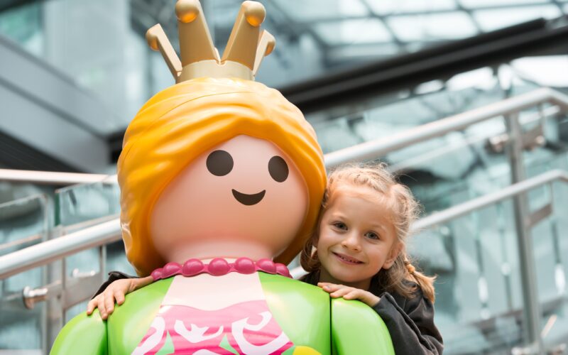 Ein junges Mädchen lächelt und umarmt eine große Playmobil-Prinzessinnenfigur mit einer Krone, drinnen in der Nähe von Glasfenstern und Metallgeländern.