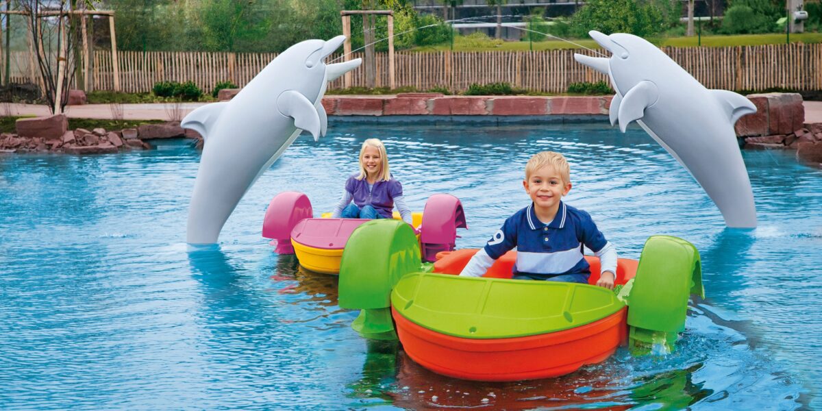 Zwei Kinder fahren mit bunten Paddelbooten in einem Pool. Im Hintergrund sind zwei große Delfinstatuen zu sehen. Hinter dem Pool sind Bäume und ein Zaun zu sehen.