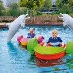 Zwei Kinder fahren mit bunten Paddelbooten in einem Pool. Im Hintergrund sind zwei große Delfinstatuen zu sehen. Hinter dem Pool sind Bäume und ein Zaun zu sehen.