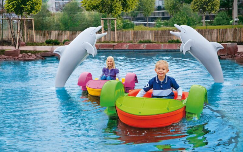 Zwei Kinder fahren mit bunten Paddelbooten in einem Pool. Im Hintergrund sind zwei große Delfinstatuen zu sehen. Hinter dem Pool sind Bäume und ein Zaun zu sehen.