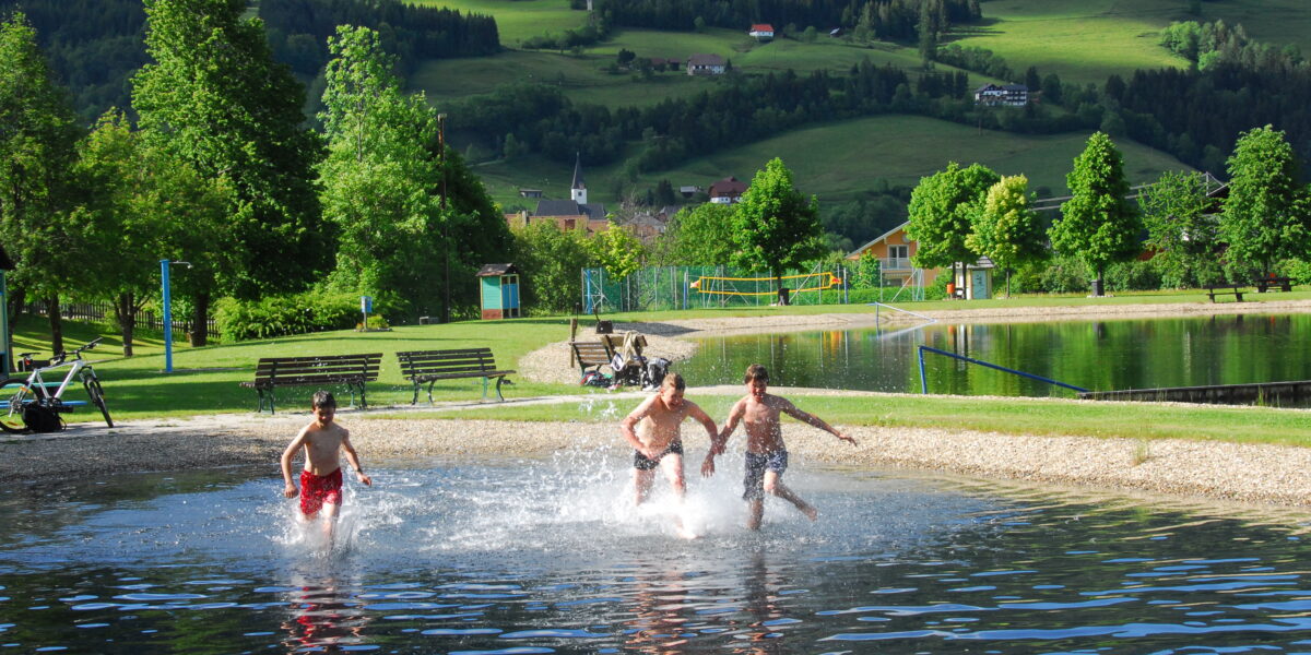 Drei Jungen rennen und planschen im seichten Wasser eines Seeufers, im Hintergrund sind grüne Hügel, Bäume und Gebäude zu sehen.