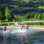 Drei Jungen rennen und planschen im seichten Wasser eines Seeufers, im Hintergrund sind grüne Hügel, Bäume und Gebäude zu sehen.