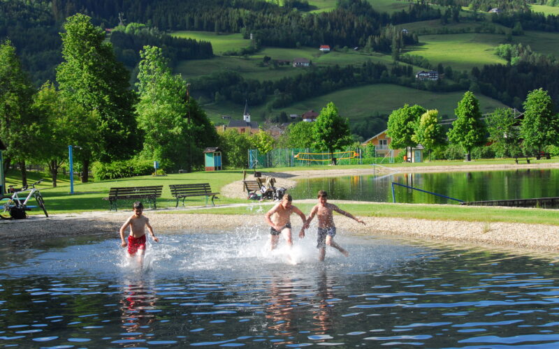 Drei Jungen rennen und planschen im seichten Wasser eines Seeufers, im Hintergrund sind grüne Hügel, Bäume und Gebäude zu sehen.
