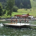 Eine Gruppe von Menschen steht und spielt auf einem Holzsteg über einem ruhigen See, mit einem Gebäude, Bäumen und grasbewachsenen Hügeln im Hintergrund.