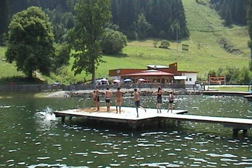 Eine Gruppe von Menschen steht und spielt auf einem Holzsteg über einem ruhigen See, mit einem Gebäude, Bäumen und grasbewachsenen Hügeln im Hintergrund.