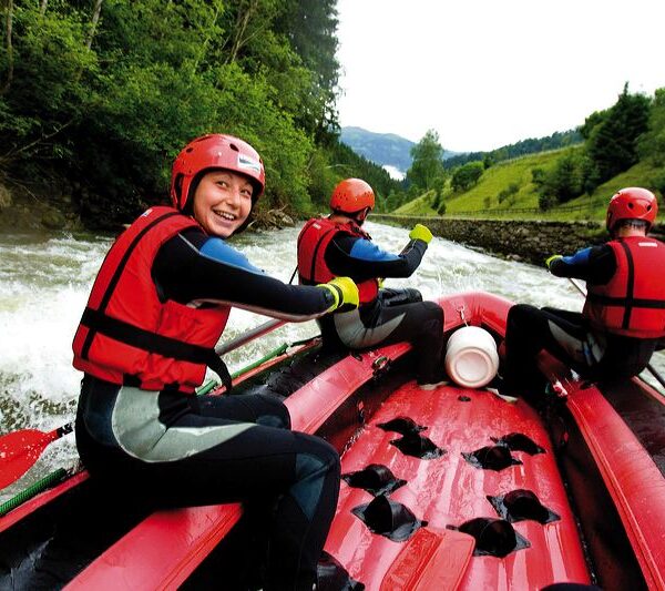 Drei Menschen mit roten Helmen und Schwimmwesten paddeln auf einem roten Schlauchboot einen von Bäumen und Hügeln umgebenen Fluss hinunter.