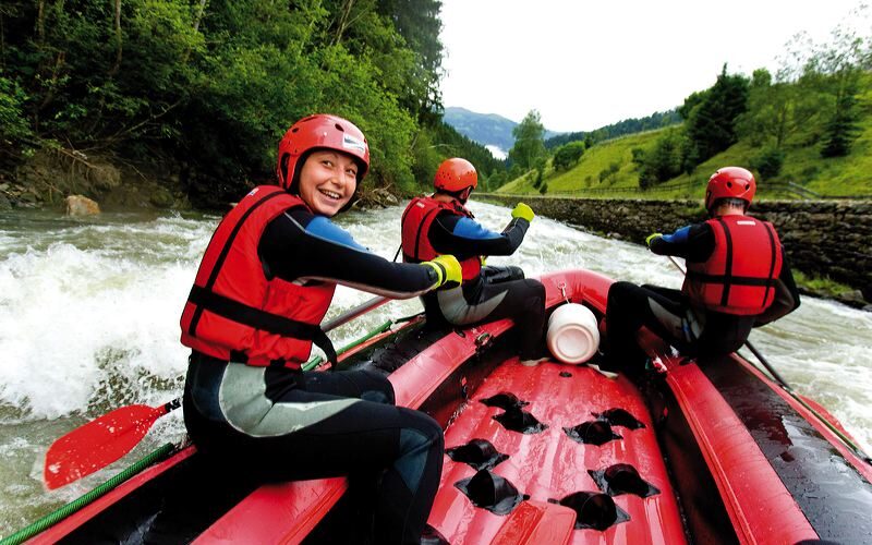 Drei Menschen mit roten Helmen und Schwimmwesten paddeln auf einem roten Schlauchboot einen von Bäumen und Hügeln umgebenen Fluss hinunter.