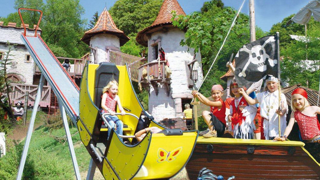 Ein Kind fährt auf einer kleinen gelben Rutsche neben einem Piratenschiff mit kostümierten Kindern vor einem skurrilen, schlossähnlichen Gebäude.