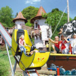 Ein Kind fährt auf einer kleinen gelben Rutsche neben einem Piratenschiff mit kostümierten Kindern vor einem skurrilen, schlossähnlichen Gebäude.