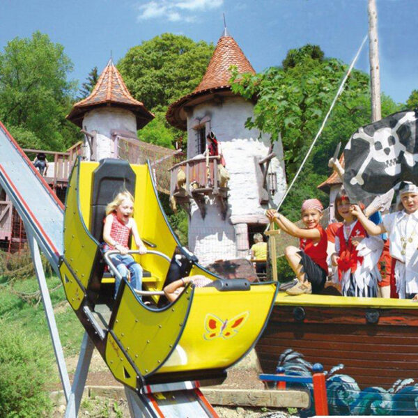 Ein Kind fährt auf einer kleinen gelben Rutsche neben einem Piratenschiff mit kostümierten Kindern vor einem skurrilen, schlossähnlichen Gebäude.