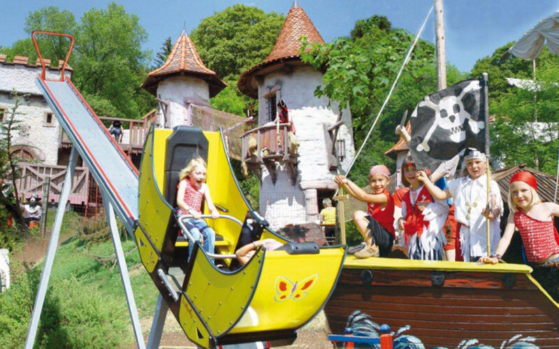 Ein Kind fährt auf einer kleinen gelben Rutsche neben einem Piratenschiff mit kostümierten Kindern vor einem skurrilen, schlossähnlichen Gebäude.