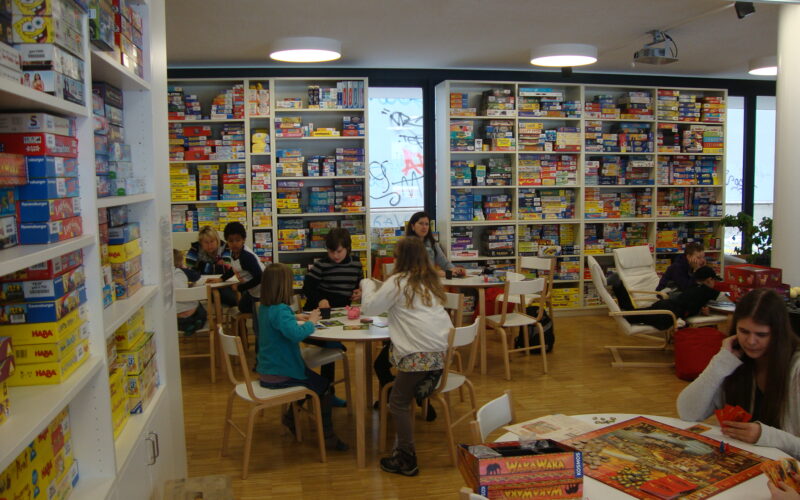 Menschen sitzen an Tischen und spielen Brettspiele in einem hell erleuchteten Raum, der mit Regalen voller verschiedener Brettspiele gefüllt ist.