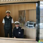 Zwei uniformierte Männer, einer stehend, der andere sitzend, befinden sich in einer hölzernen Bürokabine mit der Aufschrift "Hafenmeister", in der im Hintergrund Regale, Dokumente und ein Drucker zu sehen sind.