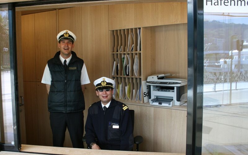 Zwei uniformierte Männer, einer stehend, der andere sitzend, befinden sich in einer hölzernen Bürokabine mit der Aufschrift "Hafenmeister", in der im Hintergrund Regale, Dokumente und ein Drucker zu sehen sind.