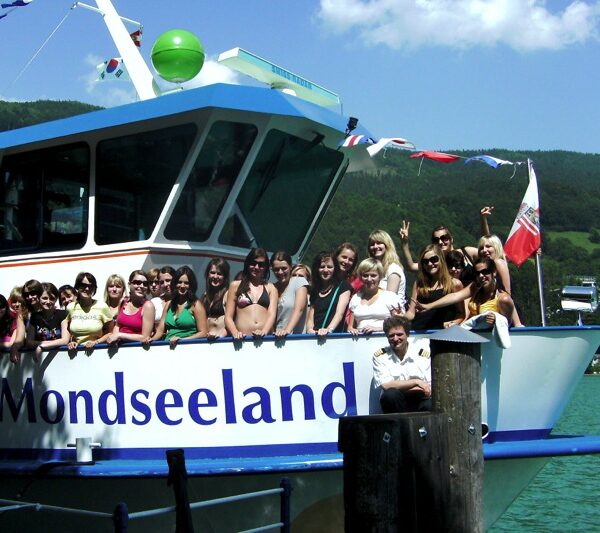 Eine Gruppe von Menschen posiert und lächelt auf dem Deck eines Bootes namens Mondseeland, das an einem See mit grünen Hügeln und Häusern im Hintergrund angedockt ist.