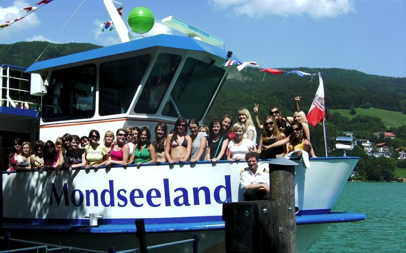 Eine Gruppe von Menschen posiert und lächelt auf dem Deck eines Bootes namens Mondseeland, das an einem See mit grünen Hügeln und Häusern im Hintergrund angedockt ist.