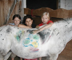 Drei Kinder stehen in einem Holzstall neben einem weißen Pferd mit bunten Farbflecken an der Seite.