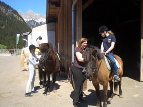 Zwei Personen bereiten Pferde vor einem Stall vor, während eine Frau einem Kind, das einen Helm trägt, beim Aufsteigen hilft. Im Hintergrund sind Berge zu sehen.