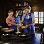 Drei Personen, zwei Erwachsene und ein Kind, tragen Schürzen in einer rustikalen Holzküche und bereiten gemeinsam an einem Herd Essen zu.