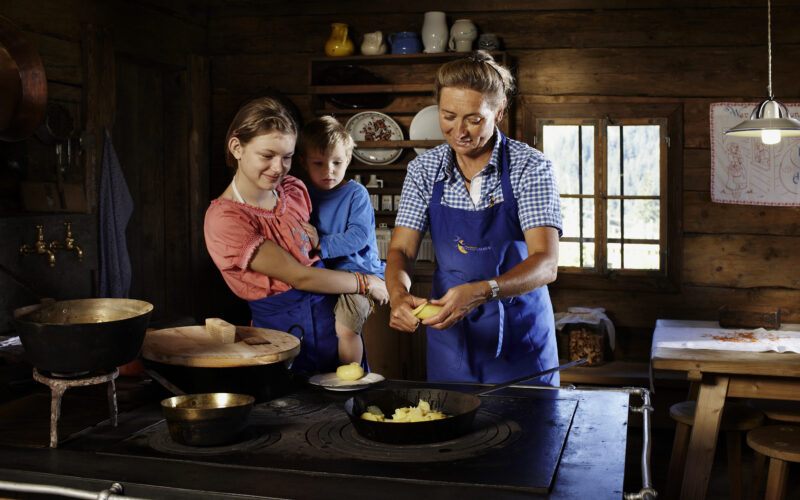 Drei Personen, zwei Erwachsene und ein Kind, tragen Schürzen in einer rustikalen Holzküche und bereiten gemeinsam an einem Herd Essen zu.