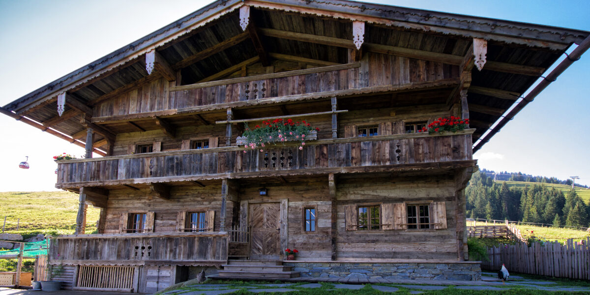 Ein rustikales Alpenchalet aus Holz mit Balkonen, Blumenkästen und einem Kreuz auf dem Dach, eingebettet in eine grasbewachsene Berglandschaft unter einem klaren blauen Himmel.