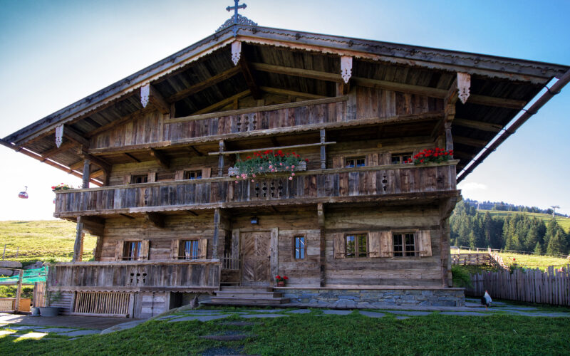 Ein rustikales Alpenchalet aus Holz mit Balkonen, Blumenkästen und einem Kreuz auf dem Dach, eingebettet in eine grasbewachsene Berglandschaft unter einem klaren blauen Himmel.