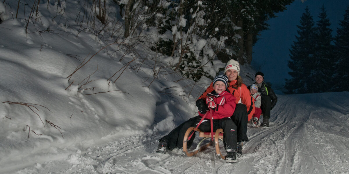 Drei winterlich gekleidete Personen fahren in der Abenddämmerung auf einem Holzschlitten einen verschneiten Hügel hinunter, mit Bäumen und Schneebänken im Hintergrund.