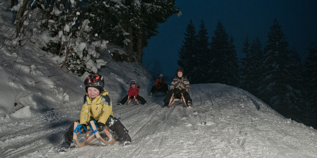 Vier Kinder rodeln nachts einen verschneiten Hügel hinunter, umgeben von schneebedeckten Bäumen, in Winterkleidung und mit Helmen.
